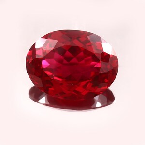 May include: A faceted, oval-shaped ruby gemstone. The gem is a deep, saturated red color with multiple facets that reflect light. The ruby sits on a reflective surface, creating a shadow.