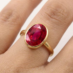 May include: A gold ring featuring a large, oval-cut, vibrant red gemstone set in a smooth, polished bezel setting. The band is thin and made of yellow gold, providing a clean and simple frame for the prominent, faceted red stone.