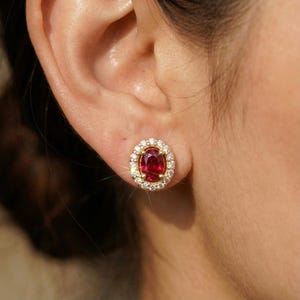 May include: Gold stud earrings featuring a central oval-cut ruby surrounded by a halo of small, round diamonds. The earrings are set in a gold metal setting, creating a classic and elegant look. The earrings are shown on an ear.