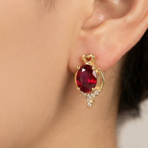 Puede incluir: Pendientes dorados con una gran piedra preciosa ovalada de color rojo intenso como punto focal. Los pendientes presentan un diseño en forma de corazón en la parte superior y pequeñas piedras de acento transparentes. Los pendientes se muestran en una oreja.