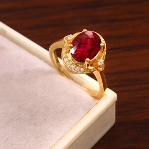 Op de afbeelding: Een gouden ring met een grote, ovaalvormige, helderrode edelsteen als middelpunt. De edelsteen is omgeven door kleinere, heldere accentstenen. De ringband en zetting zijn helder goudkleurig, wat een opvallend contrast creëert.
