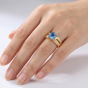Könnte beinhalten: Ein goldener Ring mit einem großen, quadratischen, hellblauen Edelstein. Der Ring hat ein geteiltes Schaftdesign mit kleinen, klaren Akzentsteinen. Der Ring wird an der Hand einer Person präsentiert.