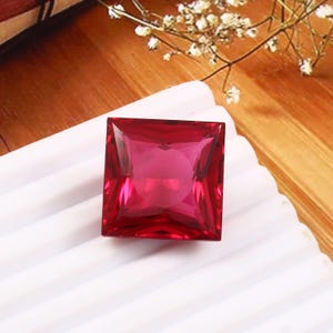 May include: A large, square, faceted, ruby-red gemstone. The gem is set on a white, textured surface, with a wooden background and delicate white flowers in the upper right corner. The gemstone is a vibrant, eye-catching focal point.