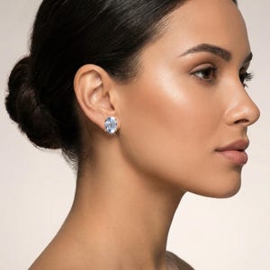 Puede incluir: Pendientes de botón de piedras preciosas ovaladas de color azul claro engastadas en oro. Los pendientes se muestran en la oreja de una persona, mostrando las brillantes piedras preciosas. Una pieza de joyería clásica.