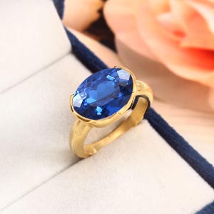 Könnte beinhalten: Ein goldener Ring mit einem großen, oval geschliffenen, leuchtend blauen Edelstein. Der Ring hat ein schlichtes, elegantes Goldband und wird in einer weißen Schmuckschatulle mit blauem Rand präsentiert. Der Hintergrund zeigt ein weiches, verschwommenes Blumenelement.