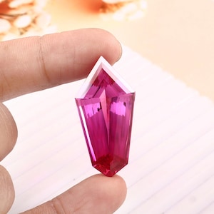 Könnte beinhalten: Ein facettierter, länglicher Edelstein in tiefem Magenta. Der Stein, gehalten zwischen zwei Fingern, hat eine spitze Spitze und eine rechteckige Basis. Die Facetten erzeugen ein Spiel von Licht und Schatten, das seinen Glanz verstärkt.