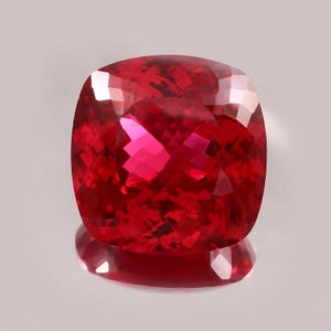 Könnte beinhalten: Ein facettierter, kissenförmiger Edelstein in tiefroter Farbe. Die Oberfläche des Edelsteins reflektiert das Licht und erzeugt ein brillantes Farb- und Funkenspiel. Der Stein ist vor einem neutralen Hintergrund platziert, der seine satte Farbe und die komplizierten Facetten hervorhebt.