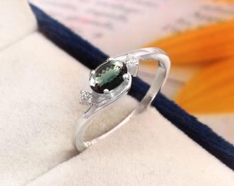 May include: A silver ring featuring an oval, dark green gemstone as the centerpiece. Two smaller, clear gemstones are set on either side of the main stone. The ring is displayed in a blue jewelry box.