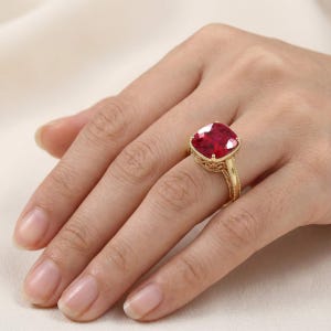 May include: A gold ring featuring a large, cushion-cut, deep red gemstone set in a decorative, filigree-style metal band. The setting includes intricate metalwork details along the sides of the band, supporting the prominent, faceted red stone.