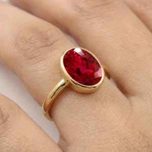 May include: A gold ring featuring a large, oval-cut, vibrant red gemstone set in a smooth, polished bezel setting. The band is thin and crafted from a polished yellow gold metal, providing a clean and simple frame for the faceted red stone.