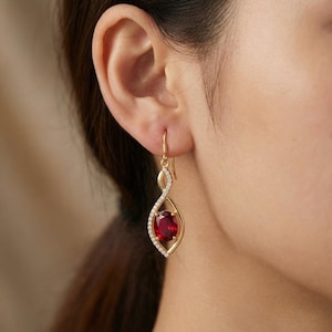 May include: Gold-tone drop earrings with a red oval gemstone center. The earrings feature a swirling design with a row of small, clear stones. The hook is a simple gold wire.