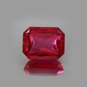 Könnte beinhalten: Ein lebendiger, rechteckig geschliffener Rubin-Edelstein. Der Edelstein hat eine tiefe, gesättigte rote Farbe mit Facetten, die das Licht reflektieren. Der Stein ist vor einem neutralgrauen Hintergrund platziert, mit einer Spiegelreflexion darunter.