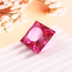 Pode incluir: Uma gema magenta radiante, de corte retangular. A gema está sobre uma superfície branca texturizada, com um fundo pêssego suave. Delicadas flores brancas são visíveis no canto superior esquerdo, adicionando elegância.
