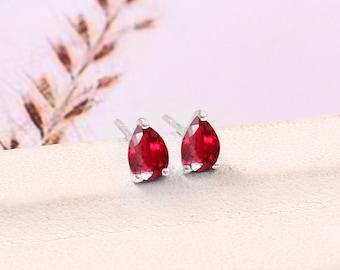May include: A pair of stud earrings featuring pear-shaped, deep red gemstones. The gemstones are set in a silver metal setting. The earrings are displayed on a white surface, with a blurred background of a plant.