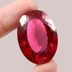 May include: A large, oval-cut gemstone in a deep red colour. The gem is held between two fingers, highlighting its polished facets and internal reflections. The stone's colour varies from a bright pink to a dark crimson.