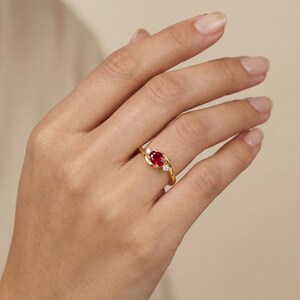 May include: A gold ring with a central oval-cut red gemstone and two smaller clear stones. The band has a flowing design. The ring is displayed on a hand. The ring's design is elegant and eye-catching.