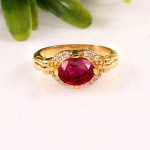 Op de afbeelding: Een gouden ring met een grote, ovaal geslepen robijn als middelpunt. De robijn is omgeven door kleinere, heldere edelstenen. De ring heeft een gedetailleerd, sierlijk ontwerp op de band.