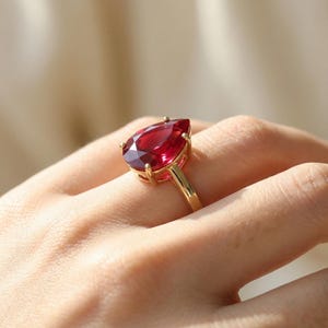 Op de afbeelding: Een gouden ring met een grote, peervormige, dieprode edelsteen. De edelsteen is gezet in een gouden band met vier klauwen. De ring wordt aan een vinger getoond.