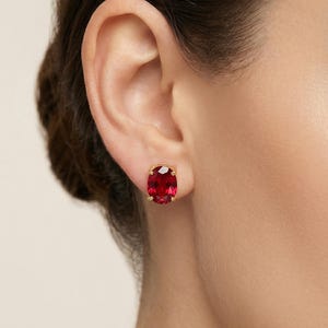Può includere: Orecchini a lobo dorati con grandi pietre preziose rosso rubino di taglio ovale. Gli orecchini sono incastonati in una semplice e classica montatura in oro. L'immagine mostra un primo piano degli orecchini su un orecchio.
