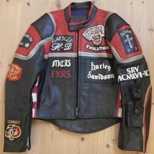 Könnte beinhalten: Schwarze und rote Motorradjacke aus Leder mit mehreren Aufnähern und Texten. Aufnäher sind u.a. "HARLEYS H-D", "EVOLUTION", "STURGIS" und "harley davidson". Die Jacke hat einen Reißverschluss und mehrere Taschen.