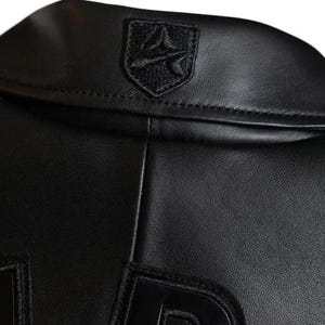 May include: Close-up of a black leather jacket collar. The jacket has a small, embroidered emblem on the upper back. The leather has a subtle texture, and the stitching is visible along the seams. The design is sleek and minimalist.