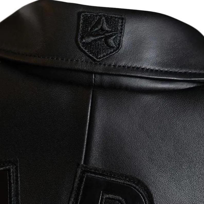 May include: Close-up of a black leather jacket collar. The jacket has a small, embroidered emblem on the upper back. The leather has a subtle texture, and the stitching is visible along the seams. The design is sleek and minimalist.