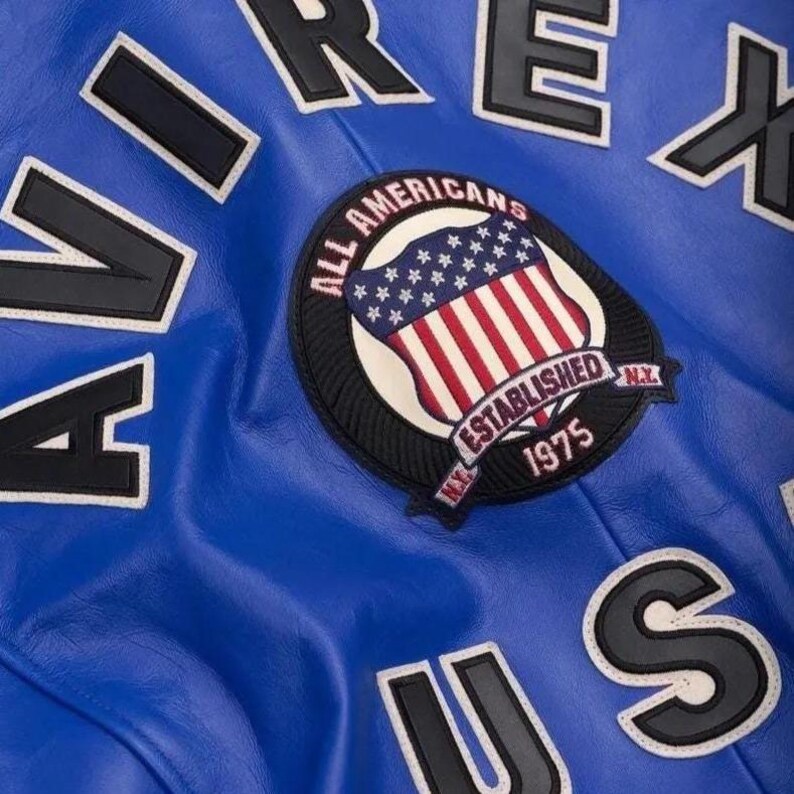 Op de afbeelding: Close-up van een blauwe leren jas met witte en zwarte letters. De jas heeft een patch met de woorden "All Americans" en "Established 1975" en een schildontwerp van de Amerikaanse vlag.