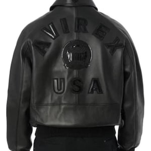 May include: Black leather jacket featuring the word "AVIREX" across the shoulders and "USA" below. The jacket has a ribbed hem and cuffs. A circular emblem is centered between the words. The jacket is a classic bomber style.