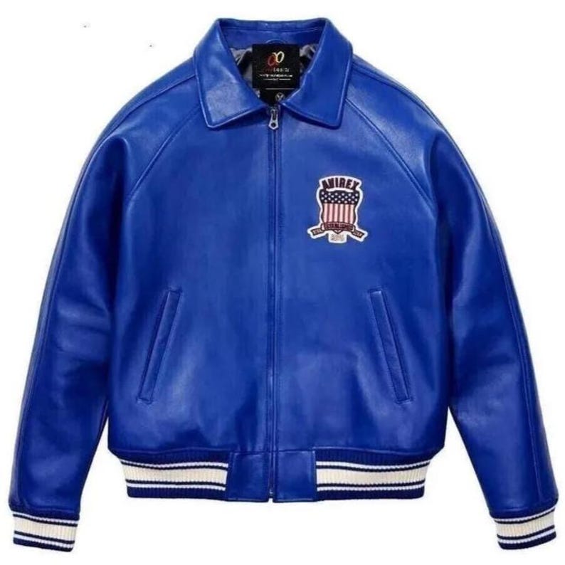 Op de afbeelding: Een helderblauwe leren jas met een klassieke kraag en een ritssluiting. De jas heeft een patch op de borst met een patriottisch ontwerp en wit en blauw gestreepte manchetten en zoom. Een stijlvol bovenkledingstuk.