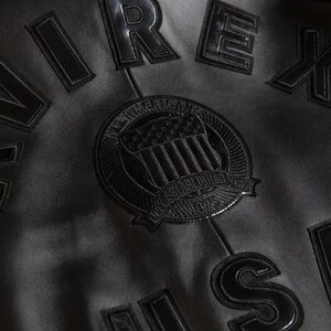 May include: Close-up of a black leather jacket with raised lettering and a circular emblem. The emblem reads "All American" and "Established 1975 NY". The jacket has the letters "REX" and "S" visible.