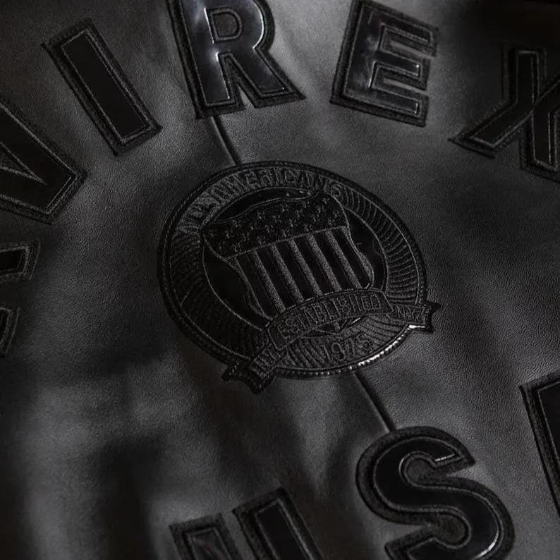 May include: Close-up of a black leather jacket with raised lettering and a circular emblem. The emblem reads "All American" and "Established 1975 NY". The jacket has the letters "REX" and "S" visible.