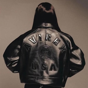 May include: Black leather jacket featuring the word "AVIREX" arched across the back, above a circular emblem and the word "USA". The jacket has a classic bomber style with a ribbed collar and cuffs. The jacket is worn by a person.