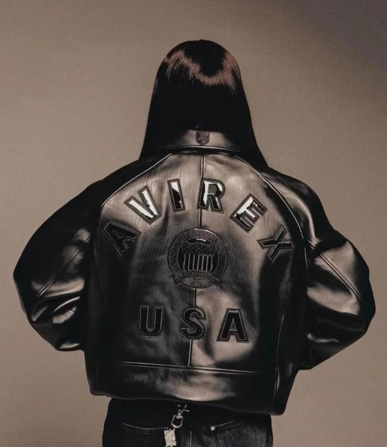 May include: Black leather jacket featuring the word "AVIREX" arched across the back, above a circular emblem and the word "USA". The jacket has a classic bomber style with a ribbed collar and cuffs. The jacket is worn by a person.