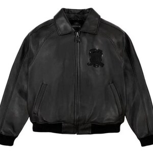 May include: Black leather bomber jacket with a collared neckline, zip closure, and ribbed cuffs and hem. Features a decorative emblem on the chest. The jacket has two front pockets.
