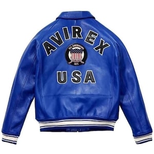 Op de afbeelding: Een felblauwe leren jas met het woord "AVIREX" in witte blokletters over de bovenrug. Het woord "USA" staat ook in het wit. Een cirkelvormige patch met een ontwerp van de Amerikaanse vlag staat tussen de woorden.