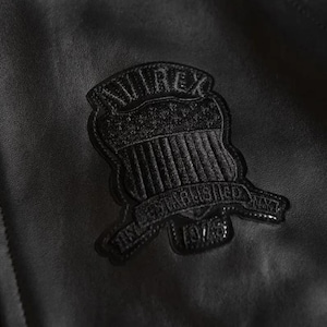 May include: Close-up of a black leather jacket with a textured patch. The patch is shield-shaped, with "AVIREX" at the top and "ESTABLISHED 1975" on a banner. The jacket has a subtle sheen, typical of leather.