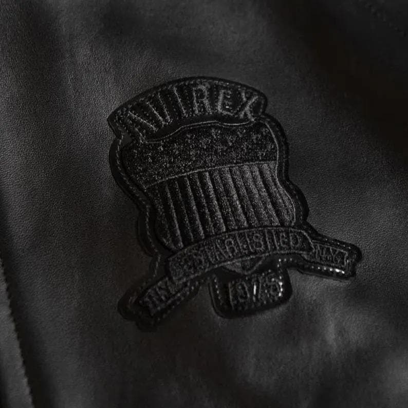 May include: Close-up of a black leather jacket with a textured patch. The patch is shield-shaped, with "AVIREX" at the top and "ESTABLISHED 1975" on a banner. The jacket has a subtle sheen, typical of leather.