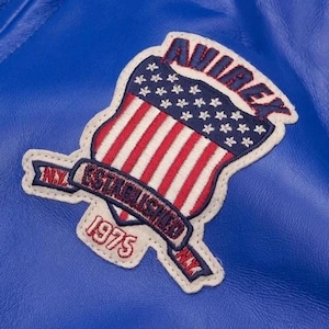 Op de afbeelding: Close-up van een blauwe leren jas met een patch. De patch heeft een schildontwerp met de Amerikaanse vlag, het woord "AVIREX" in blauw en de tekst "ESTABLISHED 1975 NY".