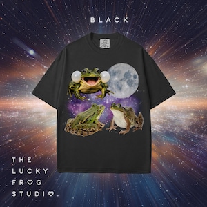 May include: Black t-shirt featuring a whimsical design of three frogs, a full moon, and a cosmic background. The shirt has the word "BLACK" at the top and "THE LUCKY FROG STUDIO" at the bottom.