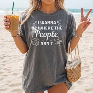May include: Gray t-shirt with the text "I Wanna Be Where The People Arn't" in white. The design includes a seashell and starfish graphic. The person is holding a drink and a straw bag on a sandy beach.