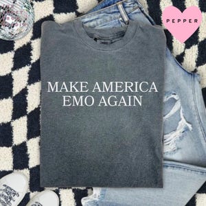 May include: A dark gray t-shirt with the text "MAKE AMERICA EMO AGAIN" in white. The shirt is folded and placed on a black and white checkered rug, with a pair of blue jeans and white sneakers in the background.