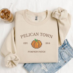 May include: A beige crewneck sweatshirt featuring the brown text PELICAN TOWN, EST. 2016, and PUMPKIN PATCH, with a central pixel art orange pumpkin graphic. The sweatshirt is paired with blue denim jeans and a glass of iced coffee.