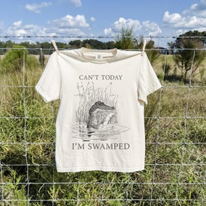 May include: Cream-colored t-shirt with the text "CAN'T TODAY I'M SWAMPED" and a black and white illustration of an alligator in water. The shirt is hanging on a clothesline outdoors.