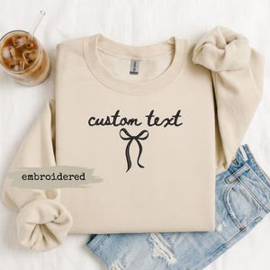 May include: A beige crewneck sweatshirt featuring black embroidery of the words custom text above a simple bow graphic. The garment has long sleeves with ribbed cuffs and a ribbed neckline. A glass of iced coffee is positioned to the top left.