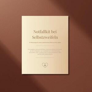Könnte beinhalten: Eine quadratische, cremefarbene Karte mit dem deutschen Text "Notfallkit bei Selbstzweifeln" ist auf einem braunen Hintergrund zentriert. Die Karte enthält zusätzlichen Text und ein herzförmiges Design. Die Beleuchtung erzeugt einen Schatteneffekt.