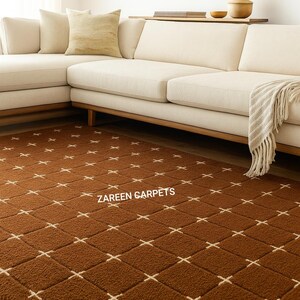 Könnte beinhalten: Ein brauner Teppich mit einem sich wiederholenden Muster aus cremefarbenen Kreuzen. Der Teppich liegt in einem Wohnzimmer unter einem cremefarbenen Sofa und Holzakzenten. Der Text "ZAREEN CARPETS" ist auf dem Teppich zu sehen.