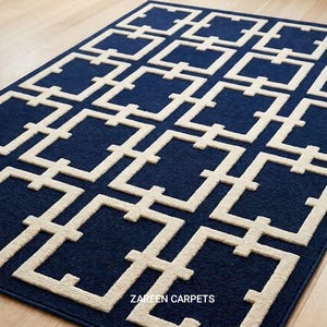 May include: A rectangular area rug featuring a deep navy blue background with a repeating cream-coloured geometric grid pattern. The text ZAREEN CARPETS is printed in white, and a small brown leather tag is attached to the bottom right corner.