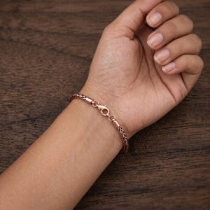 May include: A copper-toned wheat chain bracelet featuring a polished finish and a secure lobster claw clasp. The bracelet is designed with a woven, braided texture that creates a continuous, flexible link pattern around the wrist area.