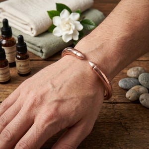 May include: A copper-colored cuff bracelet with pointed ends is worn on a wrist. The bracelet is the focal point, with a wooden surface and essential oil bottles in the background. A white flower and folded towels add to the spa-like aesthetic.