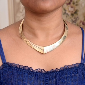 Puede incluir: Un collar rígido de metal en tono dorado pulido con un diseño liso en forma de V que descansa en la base del cuello. El collar tiene una estructura de banda curva y sólida con una superficie reflectante y un detalle central puntiagudo.
